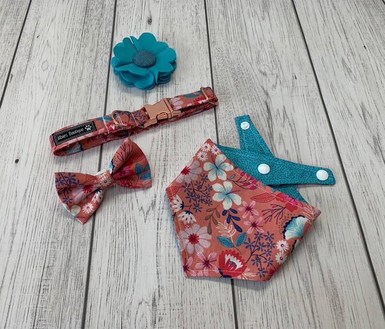 May include: A floral dog collar, bow tie, and bandana set in shades of coral, blue, and white. A matching teal felt flower is also included. The collar has a rose gold buckle and the brand name "Albies Boutique" printed on it.