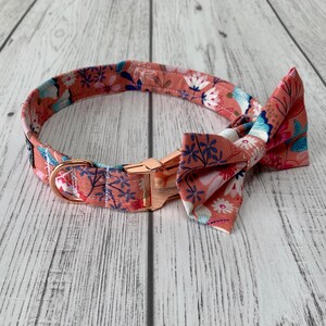 May include: A floral dog collar and bow tie set. The collar and bow tie feature a peach-colored fabric with a repeating pattern of white, blue, and navy flowers. The collar has a rose gold buckle and hardware.