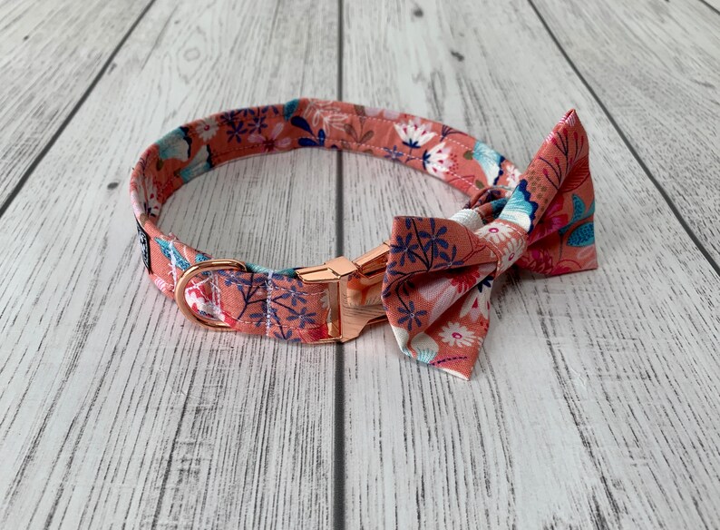 May include: A floral dog collar and bow tie set. The collar and bow tie feature a peach-colored fabric with a repeating pattern of white, blue, and navy flowers. The collar has a rose gold buckle and hardware.