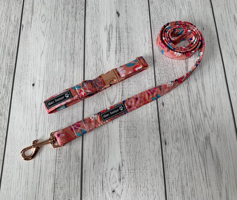 May include: A floral dog collar and leash set. The set features a peach-colored background with a colorful floral pattern. The collar has a rose gold buckle, and the leash has a matching rose gold clasp. The brand name "Abies Boutique" is printed on the collar and leash.