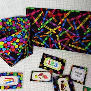 May include: A set of colorful fabric pouches with a crayon pattern. The pouches are filled with small fabric squares with different designs, including numbers, a book, and the words "Happy Day".