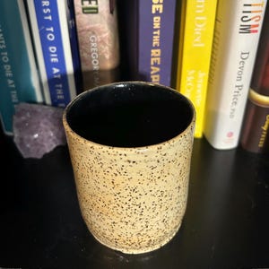 Puede incluir: Un vaso de cerámica moteada con interior negro. El exterior es de color beige claro con pequeñas motas de color marrón oscuro. El vaso es cilíndrico y se asienta sobre una superficie oscura. Se ven libros en el fondo.