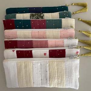 Peut inclure: Six pochettes zippées matelassées dans différentes couleurs, notamment le vert, le rose et le bordeaux. Chaque pochette a un tirette de fermeture éclair avec un pompon doré.