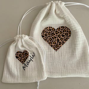 Könnte beinhalten: Zwei weiße Kordelzugbeutel mit einem herzförmigen Leopardenmuster. Der kleinere Beutel trägt den Namen "Mayli" in Schreibschrift. Die Beutel bestehen aus einem strukturierten Stoff und haben einen Kordelzugverschluss.