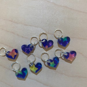 Peut inclure: Neuf marqueurs de points en forme de cœur avec des anneaux argentés. Chaque marqueur présente un motif d'étoile coloré différent sur un fond bleu foncé.