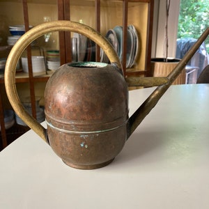 Könnte beinhalten: Eine Vintage-Gießkanne aus Kupfer mit Messinggriff und Ausguss. Die Gießkanne hat einen abgerundeten Körper und einen langen, schlanken Ausguss. Der Griff ist gebogen und die Kanne steht auf einer weißen Oberfläche.