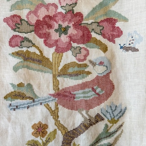 Op de afbeelding: Kruissteekborduurwerk met een vogel op een tak met bloemen en bladeren. De vogel is rood met blauwe details, en de bloemen zijn roze en rood. De achtergrond is lichtbeige.