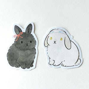 May include: Two bunny stickers. One is black with a pink bow, the other is white with a blue headband. Both have black eyes and are in a sitting position. The stickers are cut out with a white border.