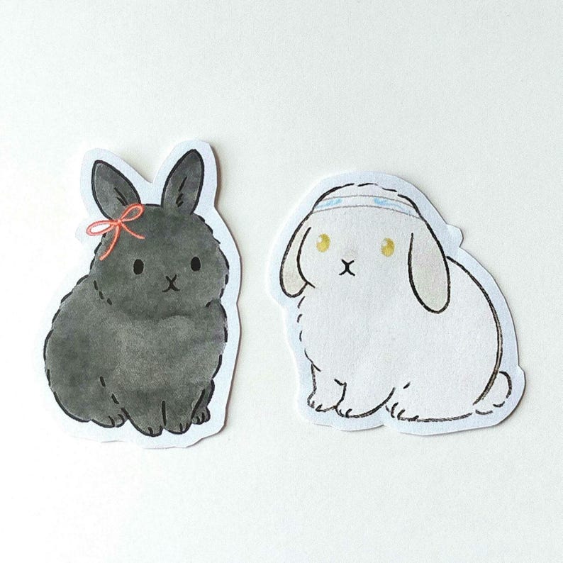 May include: Two bunny stickers. One is black with a pink bow, the other is white with a blue headband. Both have black eyes and are in a sitting position. The stickers are cut out with a white border.