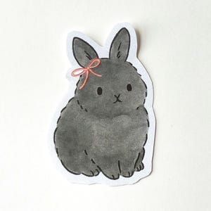 May include: A hand-drawn sticker featuring a gray bunny with a pink bow. The bunny is sitting, with its ears perked up. The sticker has a white border and is on a white background. The bunny has a cute, cartoonish appearance.