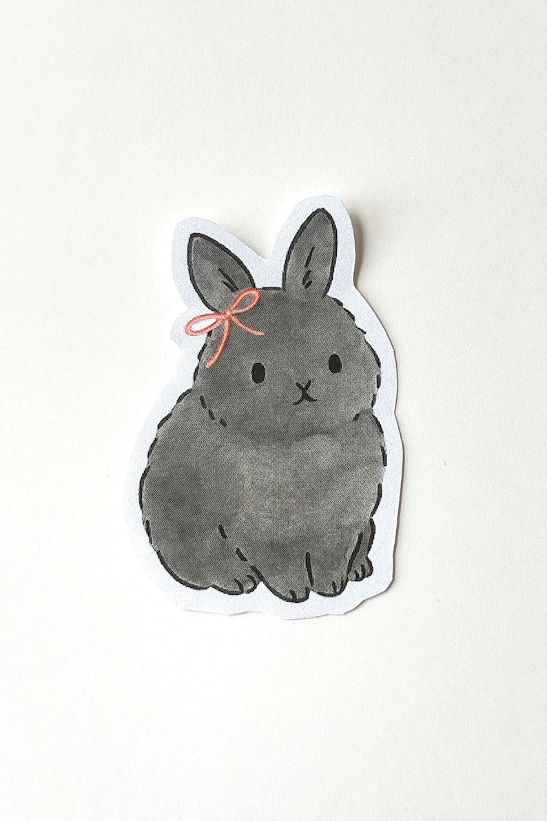 May include: A hand-drawn sticker featuring a gray bunny with a pink bow. The bunny is sitting, with its ears perked up. The sticker has a white border and is on a white background. The bunny has a cute, cartoonish appearance.