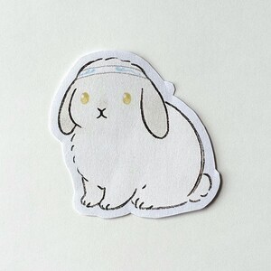 May include: A white sticker featuring a cartoon bunny with a blue headband. The bunny has yellow eyes, a small black 'x' for a mouth, and black outlines. The sticker is cut around the bunny's shape.