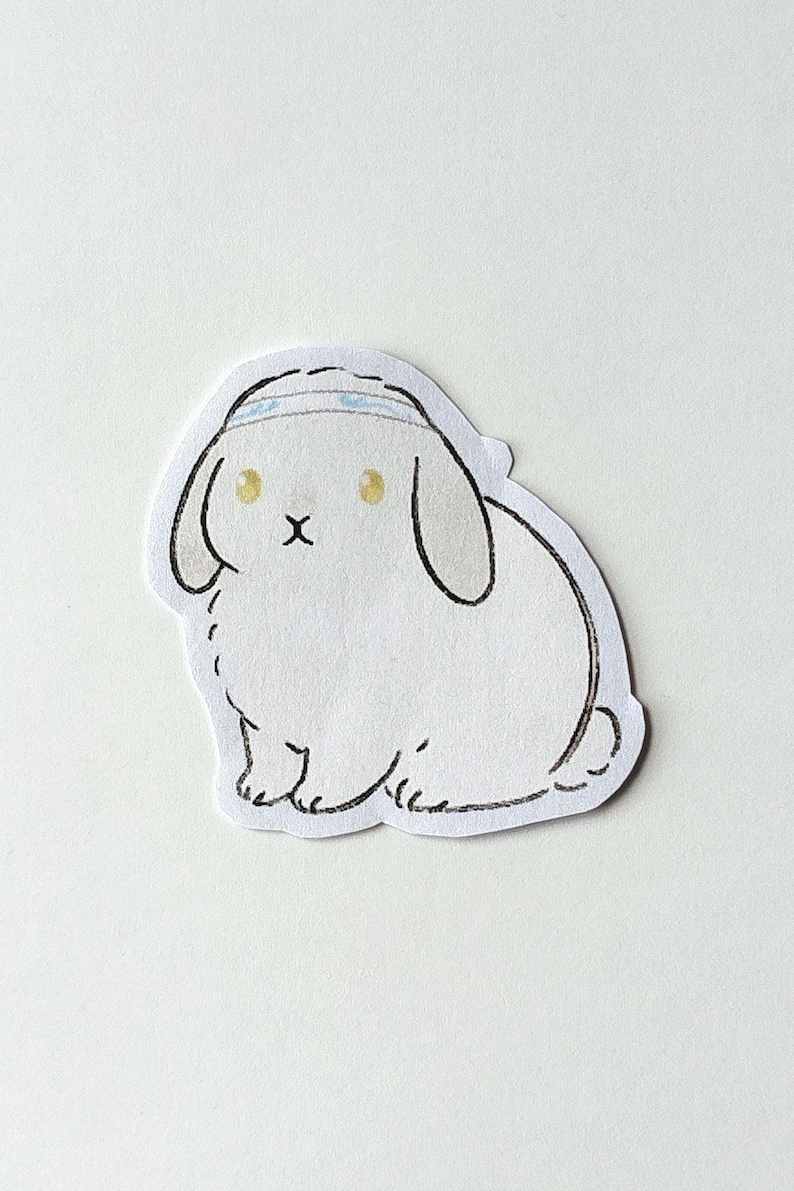 May include: A white sticker featuring a cartoon bunny with a blue headband. The bunny has yellow eyes, a small black 'x' for a mouth, and black outlines. The sticker is cut around the bunny's shape.