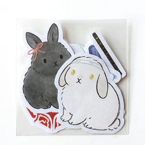 May include: A set of stickers featuring cartoon bunnies in a clear plastic sleeve. One bunny is black with a pink bow, another is white with a headband, and the third is a black flute. The stickers are on a white background.