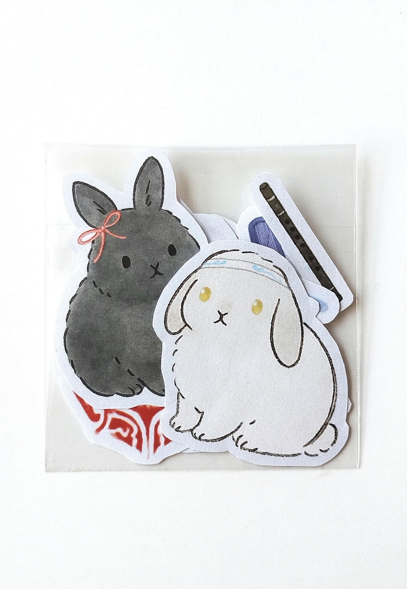 May include: A set of stickers featuring cartoon bunnies in a clear plastic sleeve. One bunny is black with a pink bow, another is white with a headband, and the third is a black flute. The stickers are on a white background.