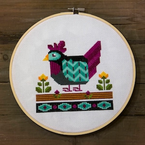 Może przedstawiać: Wzór haftu krzyżykowego przedstawiający kolorowego koguta z turkusowym, fioletowym i czarnym ciałem, stojącego na brązowej platformie z żółtymi kwiatami. Kogut jest otoczony turkusową, fioletową i czarną ramką.
