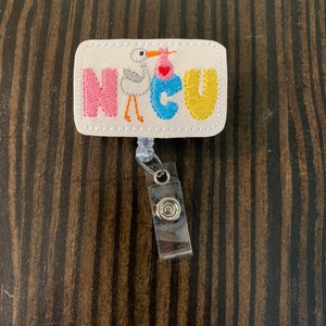 May include: A white rectangular badge with the word "NICU" in colourful embroidered letters. A white stork with a pink and blue baby in a basket is embroidered in the centre of the badge. The badge is attached to a silver retractable badge holder.