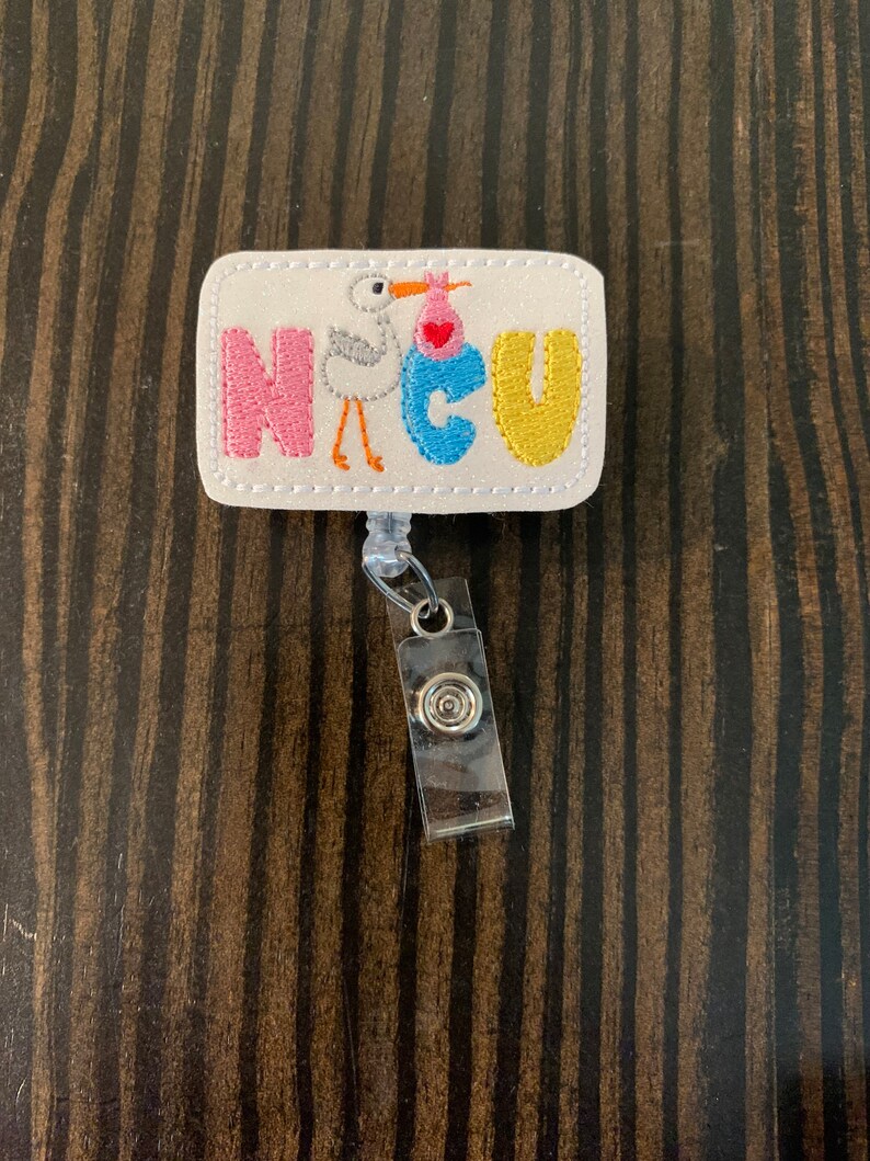 May include: A white rectangular badge with the word "NICU" in colourful embroidered letters. A white stork with a pink and blue baby in a basket is embroidered in the centre of the badge. The badge is attached to a silver retractable badge holder.