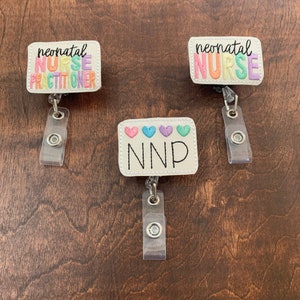 May include: Three white badge reels with colorful embroidered text. The top two reels say "neonatal nurse" and the bottom reel says "NNP" with four small hearts.