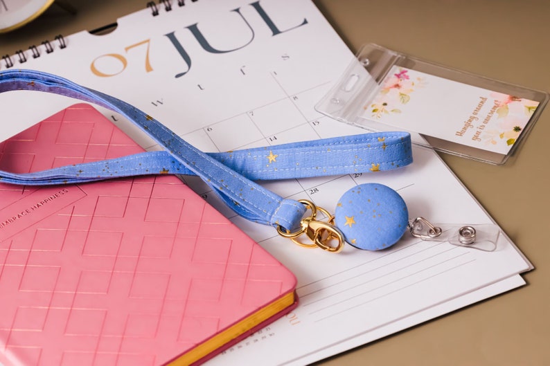 Teacher lanyard laying on desk next to a calendar. Fabric is soft cotton in baby blue with metallic gold stars. Matching badge reel and a clear ID holder is attached to lanyard.