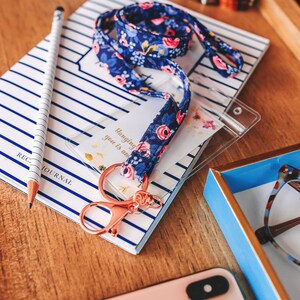 May include: A blue and white floral patterned lanyard with a rose gold key chain and a clear plastic ID badge holder. The lanyard is on top of a striped notebook with the text "REG. JOURNAL" printed on it. The text on the ID badge holder reads "Hanging with you is an adventure."