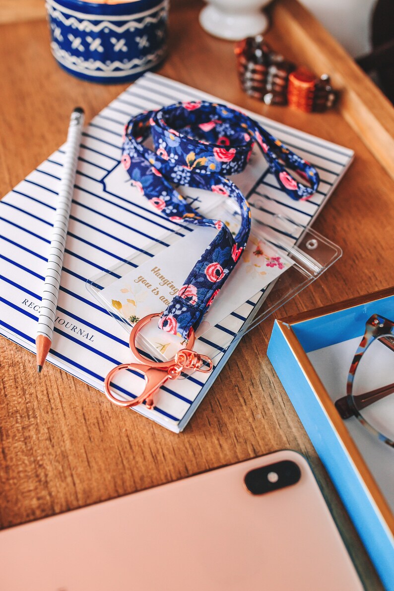 May include: A blue and white floral patterned lanyard with a rose gold key chain and a clear plastic ID badge holder. The lanyard is on top of a striped notebook with the text "REG. JOURNAL" printed on it. The text on the ID badge holder reads "Hanging with you is an adventure."