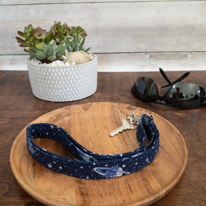 May include: A blue and white lanyard with a space theme design, featuring planets and stars. The lanyard is on a wooden tray with a key and sunglasses.