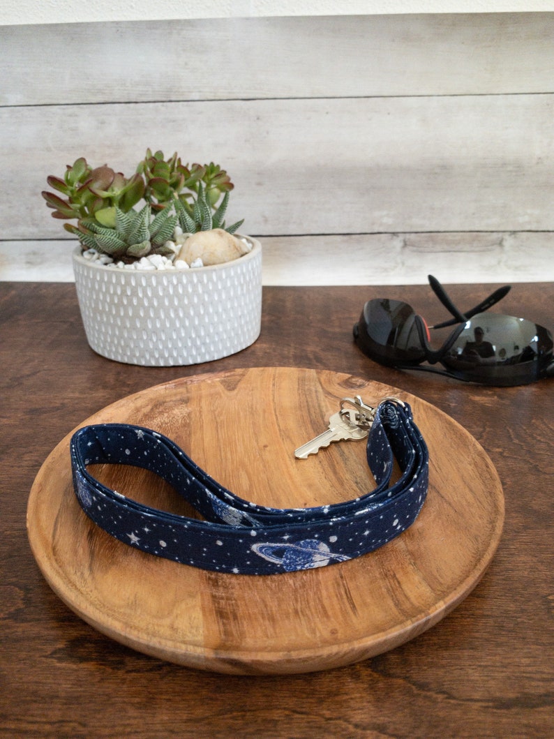 May include: A blue and white lanyard with a space theme design, featuring planets and stars. The lanyard is on a wooden tray with a key and sunglasses.