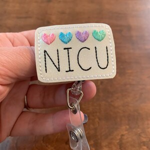 May include: A rectangular badge reel with the word "NICU" in black lettering. Above the text are four embroidered hearts in pink, blue, purple, and green. The badge reel is white with a glittery finish and has a clear plastic clip.