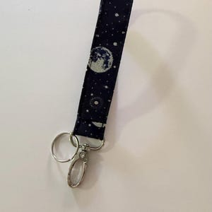 May include: A navy blue fabric wristlet key fob with a celestial design. The pattern includes a large moon and scattered white stars. It has a silver-colored metal clasp and key ring.