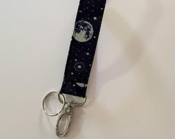 May include: A navy blue fabric wristlet key fob with a celestial design. The pattern includes a large moon and scattered white stars. It has a silver-colored metal clasp and key ring.