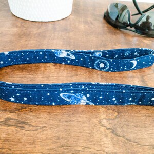 May include: A blue and white fabric key chain with a space theme. The key chain features planets, stars, and a crescent moon.