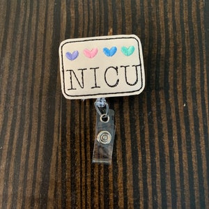 May include: A white fabric badge with four embroidered hearts in blue, pink, and teal. The badge reads "NICU" in black embroidery. The badge is attached to a clear plastic retractable badge holder.