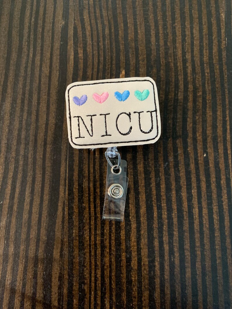 May include: A white fabric badge with four embroidered hearts in blue, pink, and teal. The badge reads "NICU" in black embroidery. The badge is attached to a clear plastic retractable badge holder.
