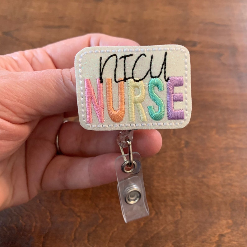 May include: A rectangular badge reel with the embroidered text "NICU NURSE" in black and pastel colors. The badge has a glittery, off-white background and a clear plastic clip.