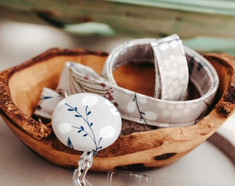 May include: A gray and white floral patterned lanyard with a matching button and a clear plastic badge holder. The lanyard is coiled in a wooden bowl.