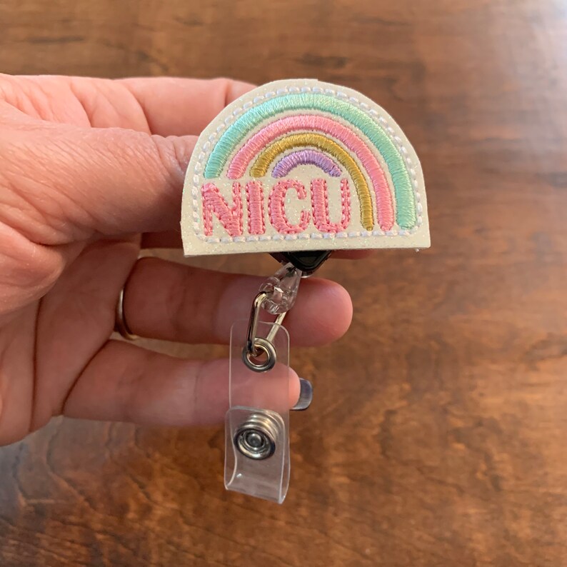 May include: A badge reel with a pastel rainbow design and the word "NICU" embroidered in pink. The badge reel has a clear plastic clip and a retractable cord.