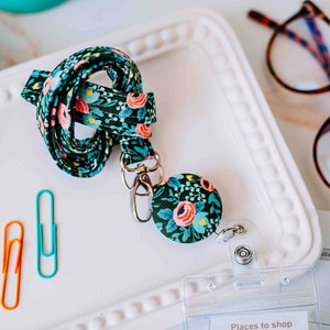 May include: A floral patterned lanyard with a retractable badge holder. The lanyard is green with pink and yellow flowers. The badge holder is a round, silver-colored clip with a floral pattern.
