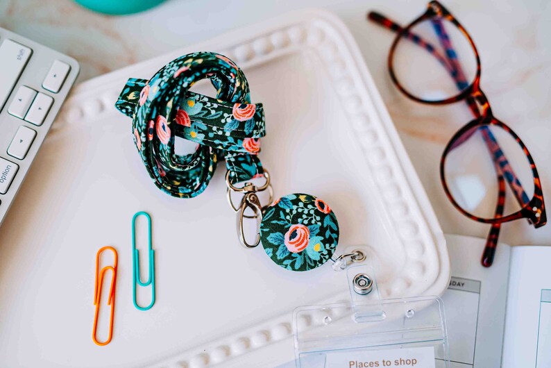 May include: A floral patterned lanyard with a retractable badge holder. The lanyard is green with pink and yellow flowers. The badge holder is a round, silver-colored clip with a floral pattern.