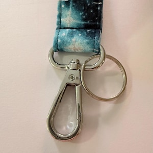 May include: A lanyard with a galaxy print in shades of blue, white, and black. The lanyard is attached to a silver-colored metal swivel clasp and a split ring. The fabric has a celestial pattern.