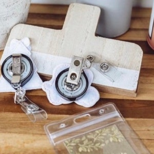 May include: Two white fabric badge holders with black retractable badge reels attached. The holders are shaped like a Christmas tree and a flower. A clear plastic badge holder is also visible in the image.
