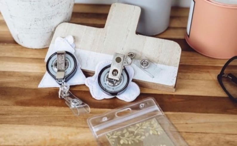 May include: Two white fabric badge holders with black retractable badge reels attached. The holders are shaped like a Christmas tree and a flower. A clear plastic badge holder is also visible in the image.