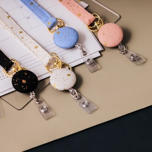 Fabric lanyards with matching badge reels laying on a desk. Black fabric with gold stars and gold hardware. White fabric with gold stars and gold hardware. Blue fabric with gold stars and gold hardware. Pink fabric with gold stars and gold hardware.