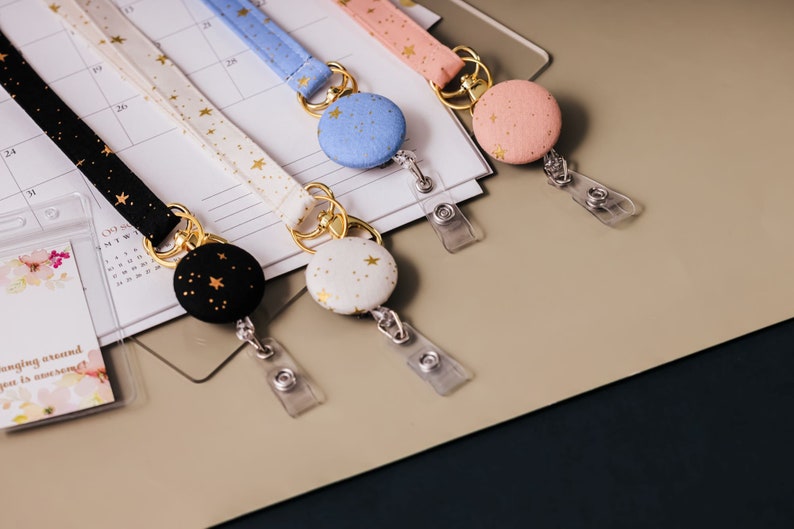 Fabric lanyards with matching badge reels laying on a desk. Black fabric with gold stars and gold hardware. White fabric with gold stars and gold hardware. Blue fabric with gold stars and gold hardware. Pink fabric with gold stars and gold hardware.