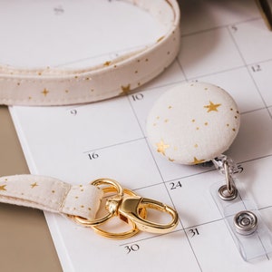 Lanyard with a matching badge reel laying on desk. Fabric is a soft white with metallic gold accents of stars and gold hardware.