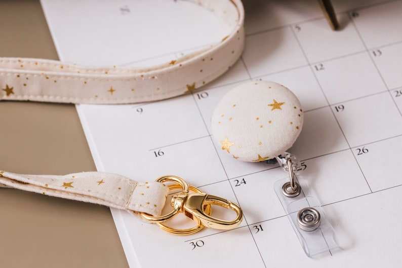 Lanyard with a matching badge reel laying on desk. Fabric is a soft white with metallic gold accents of stars and gold hardware.