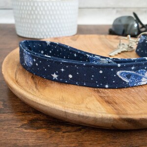 May include: A blue and white fabric key chain with a space theme. The key chain features a pattern of stars, planets, and a galaxy design.