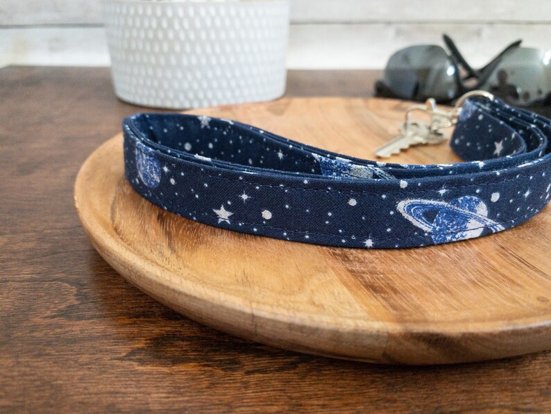 May include: A blue and white fabric key chain with a space theme. The key chain features a pattern of stars, planets, and a galaxy design.