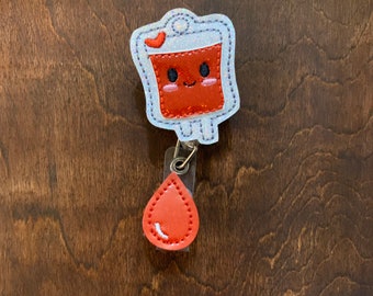 May include: A cute, red blood bag badge reel with a smiling face and a heart on top. The badge reel has a glittery white border and a red teardrop charm attached. The background is a dark brown wooden surface.