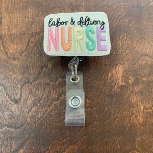May include: A white badge holder with a colorful embroidered badge that says "labor & delivery NURSE". The badge is attached to a clear plastic retractable badge reel.